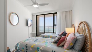 A hotel room with two beds, a ceiling fan, a TV, and a balcony with a view of the sea.
