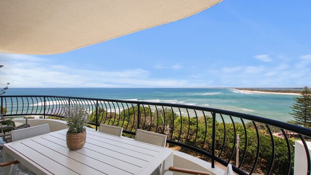 A balcony with a white table, chairs, and a potted plant. It overlooks a beach with the ocean and a clear sky.