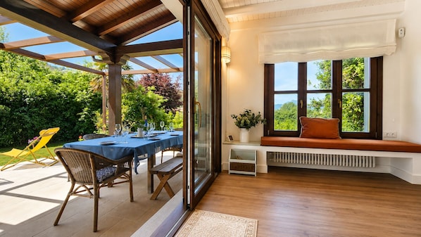 A covered patio with a table set for dining, a bench with a cushion, and a view of greenery through large windows.