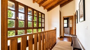 Un pasillo con barandillas de madera, ventanas amplias y vistas al exterior con vegetación.