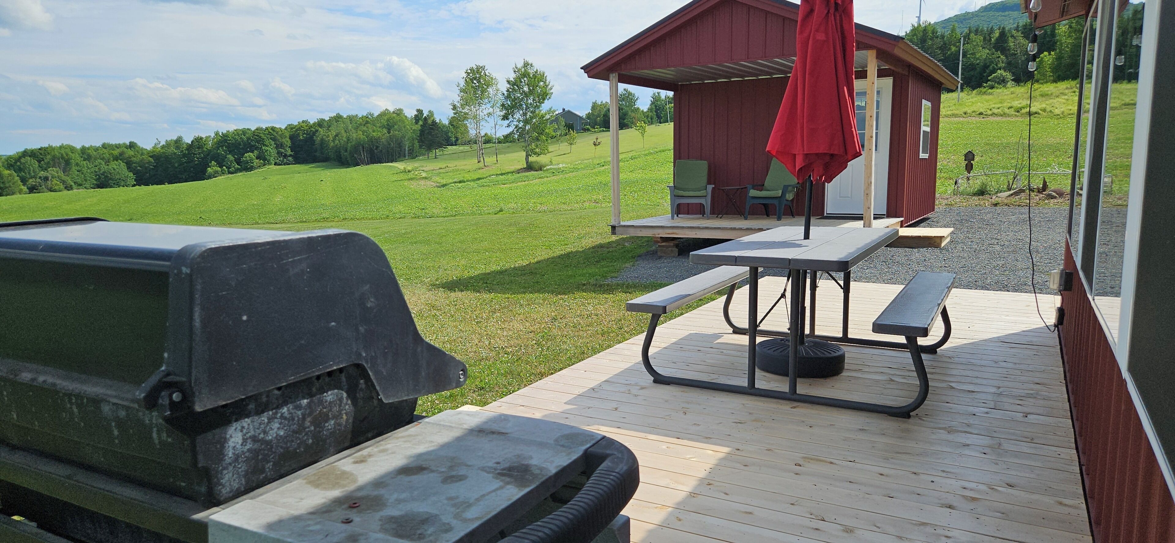 Une terrasse avec des tables de pique-nique et un barbecue, donnant sur un champ verdoyant et un bâtiment rouge.