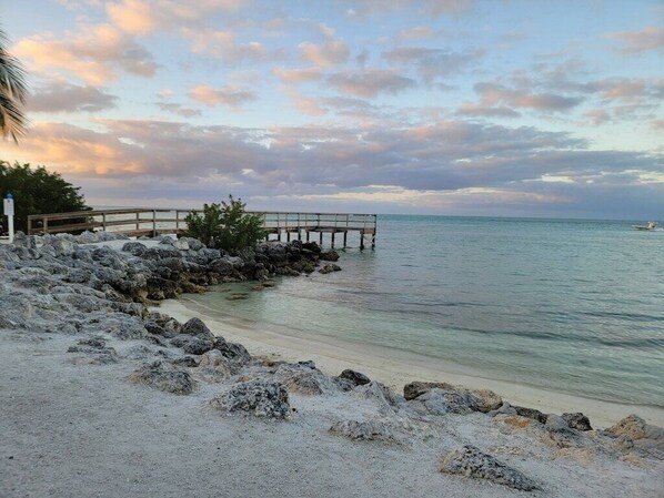 A beach with a wooden pier extending into calm waters, surrounded by rocks and a clear sky.
