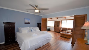 A bedroom with a large bed, wooden floor, ceiling fan, and a view into the living area.