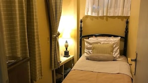 Une petite chambre à coucher accueillante, avec un lit simple, une table de chevet avec une lampe et une fenêtre ornée de rideaux blancs.