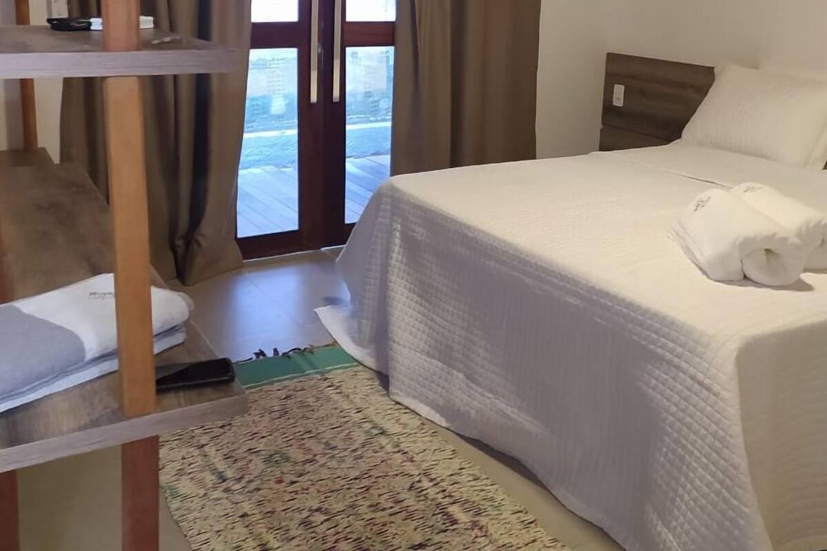 Quarto com cama, prateleira de madeira, janela com cortinas e vista para o exterior.