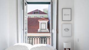 Ein Schlafzimmer mit Bett, Kissen, einem Nachttisch, einer Lampe und Blick auf Gebäude durch eine offene Tür.