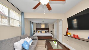 Une chambre d’hôtel avec un canapé gris, une table basse en bois, un lit avec une tête de lit, un ventilateur de plafond et un téléviseur fixé au mur.
