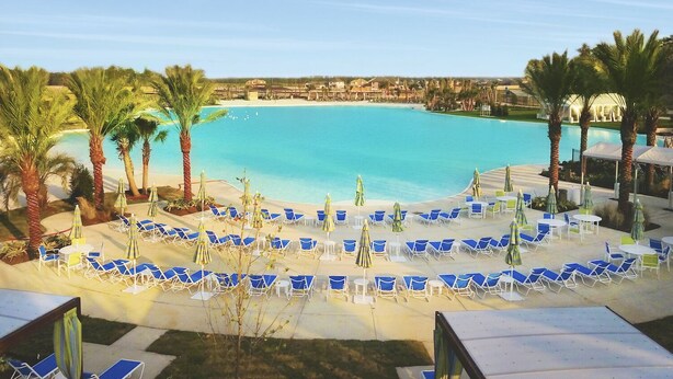 Une piscine d’hôtel avec des chaises et des parasols bleus, entourée de palmiers et offrant une vue dégagée sur l’océan.