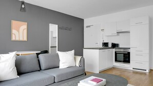 Een moderne woonkamer met een grijze bank, een bijzettafeltje met boeken en een keuken met witte kasten en een inbouwoven.