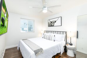 Une chambre à coucher comprenant un lit, un ventilateur de plafond, un téléviseur fixé au mur, une fenêtre avec des stores, un tableau encadré, une table de chevet avec une lampe et une plante sur la table de chevet.