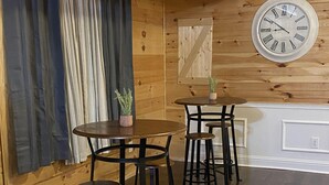 A wooden interior with a round table and four stools, a clock on the wall, and curtains on the window.