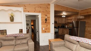 A cozy living room with two sofas and a coffee table, wooden panel walls, and a kitchen in the background.