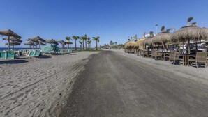 A beach with rows of thatched umbrellas and beach chairs, palm trees, and a clear sky. - A&n House of the Sun . (Torre del Mar)