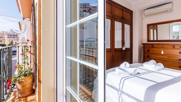 A balcony with a potted plant and a view of the cityscape. The room has a wooden dresser, a bed with white linens, and an air conditioning unit on the wall. - A&n House of the Sun . (Torre del Mar)