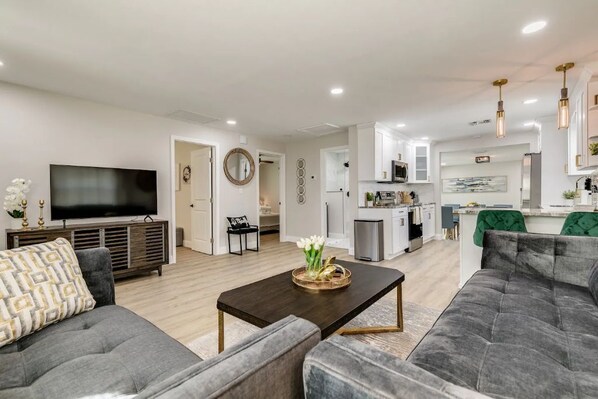 A modern living room with a flat-screen TV, grey sofas, a coffee table, and a kitchen in the background.