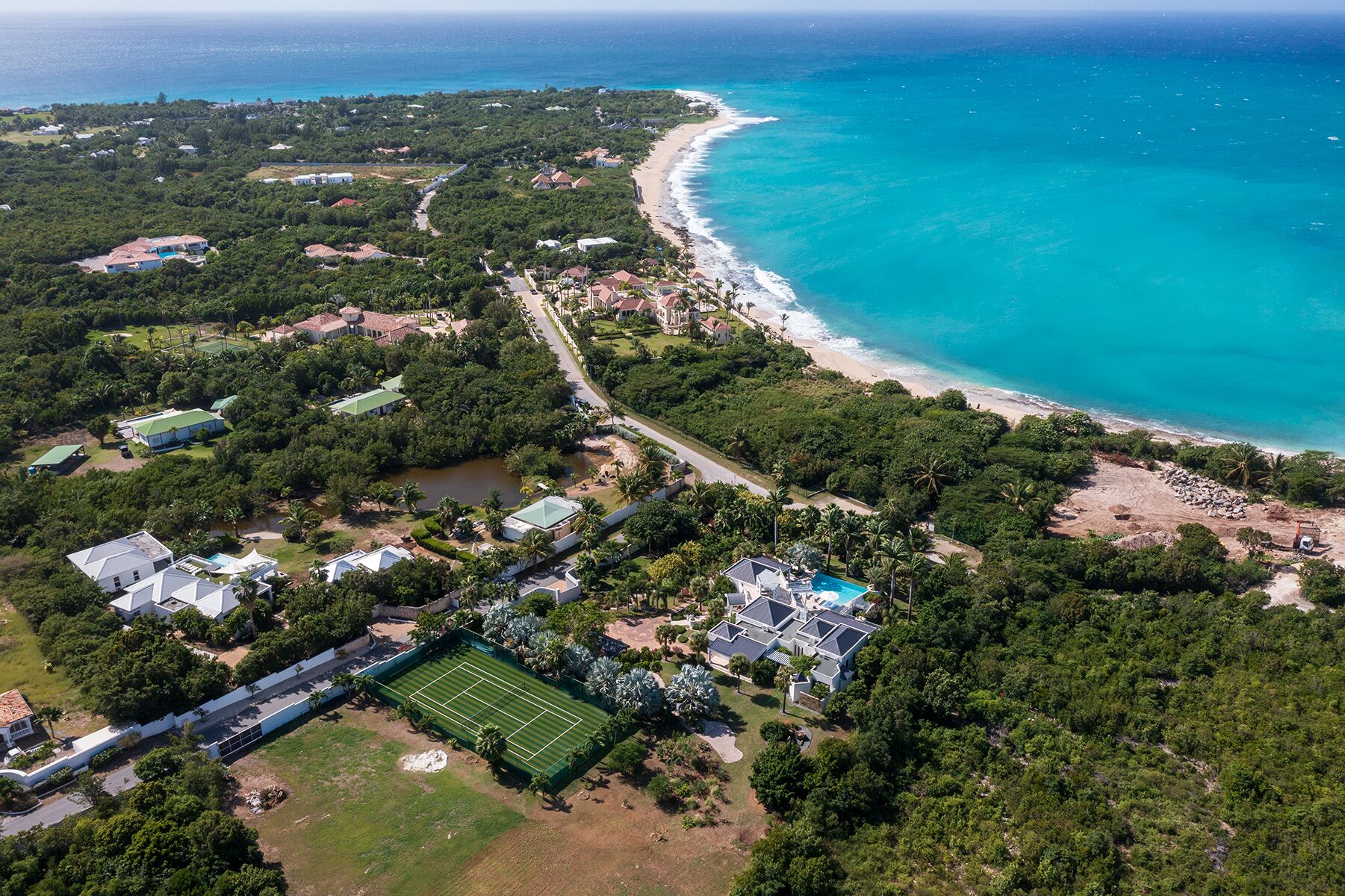 An aerial view of a coastal property with a beach, tennis court, and residential buildings.