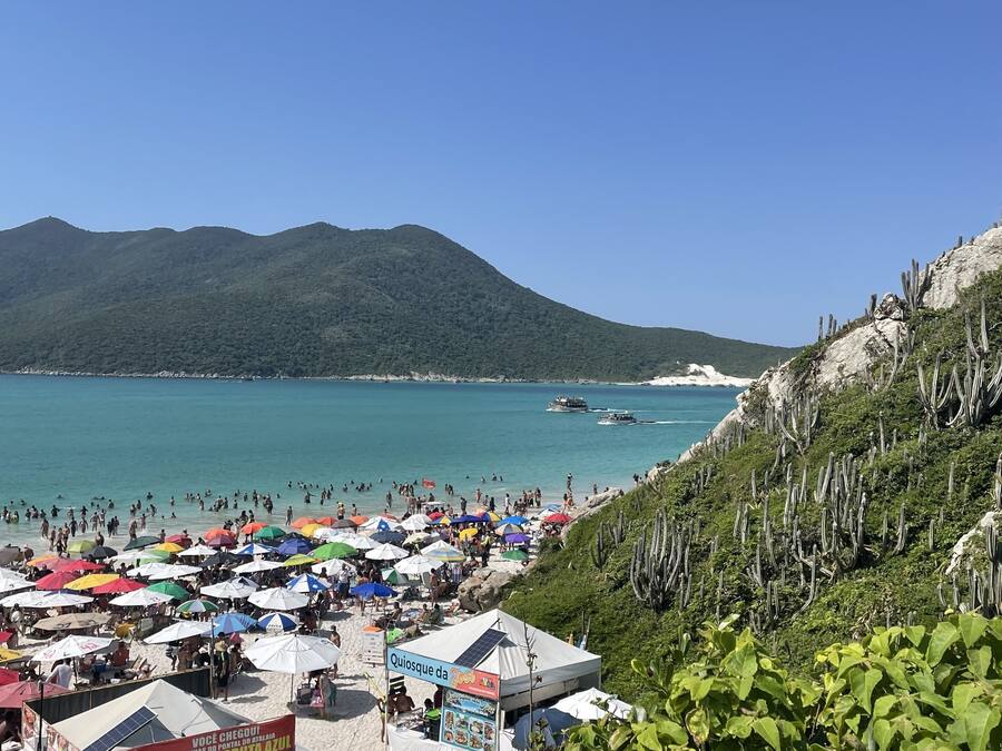 A beach with numerous colorful umbrellas, a clear blue sky, and a mountainous backdrop.