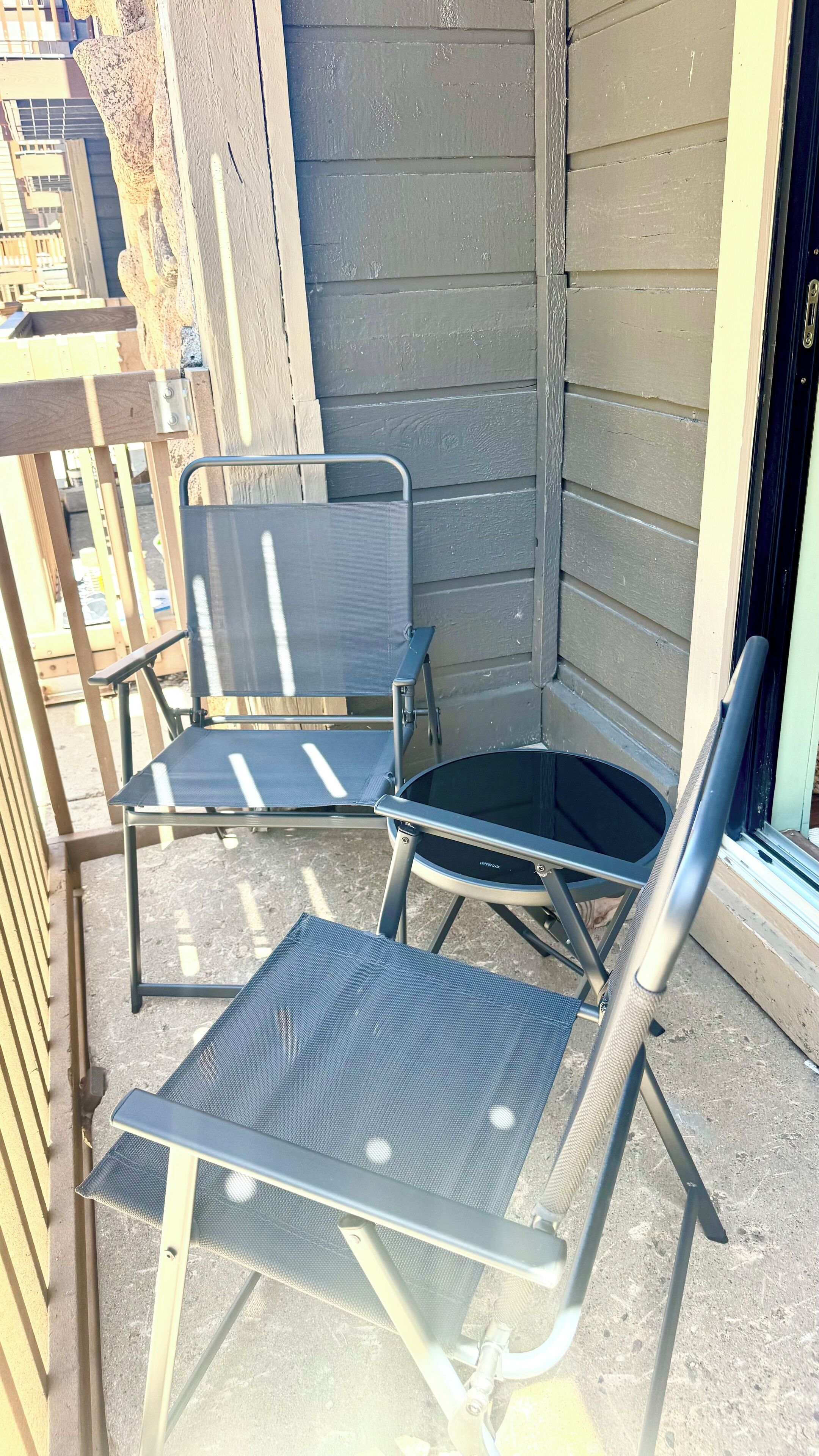 Een hoekje met twee metalen stoelen en een kleine tafel, een houten hek en een gebouw met beige bekleding.