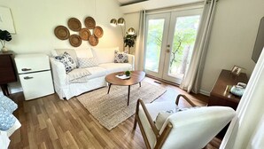 Ein modernes Wohnzimmer mit einem weißen Sofa, einem Couchtisch und Holzboden. Es gibt eine kleine weiße Kommode und eine Pflanze auf dem Fensterbrett.