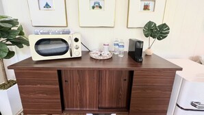 Ein Holztisch mit einer Mikrowelle, einer Snackbox, zwei Wasserflaschen und einer Kaffeemaschine. An der Wand hinter dem Tisch hängen gerahmte Bilder.