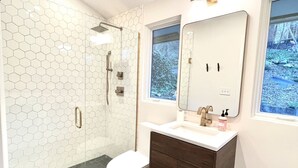 Ein modernes Badezimmer mit Wand aus sechseckigen Fliesen, einer begehbaren Dusche, einer Toilette, einem Waschtisch mit Spiegel und einem Fenster mit Blick auf Bäume.