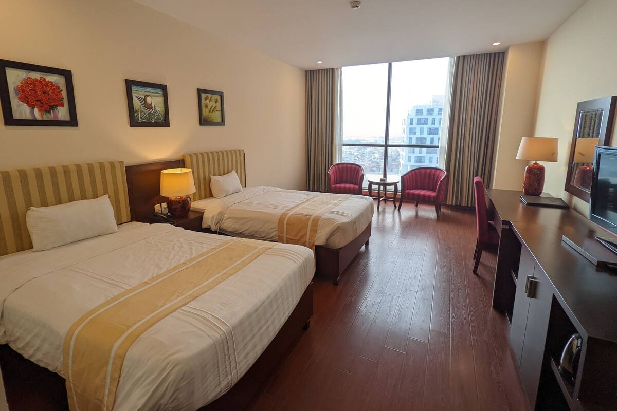 A hotel room with two beds, a desk with a TV, a chair, and a view of the city through large windows.