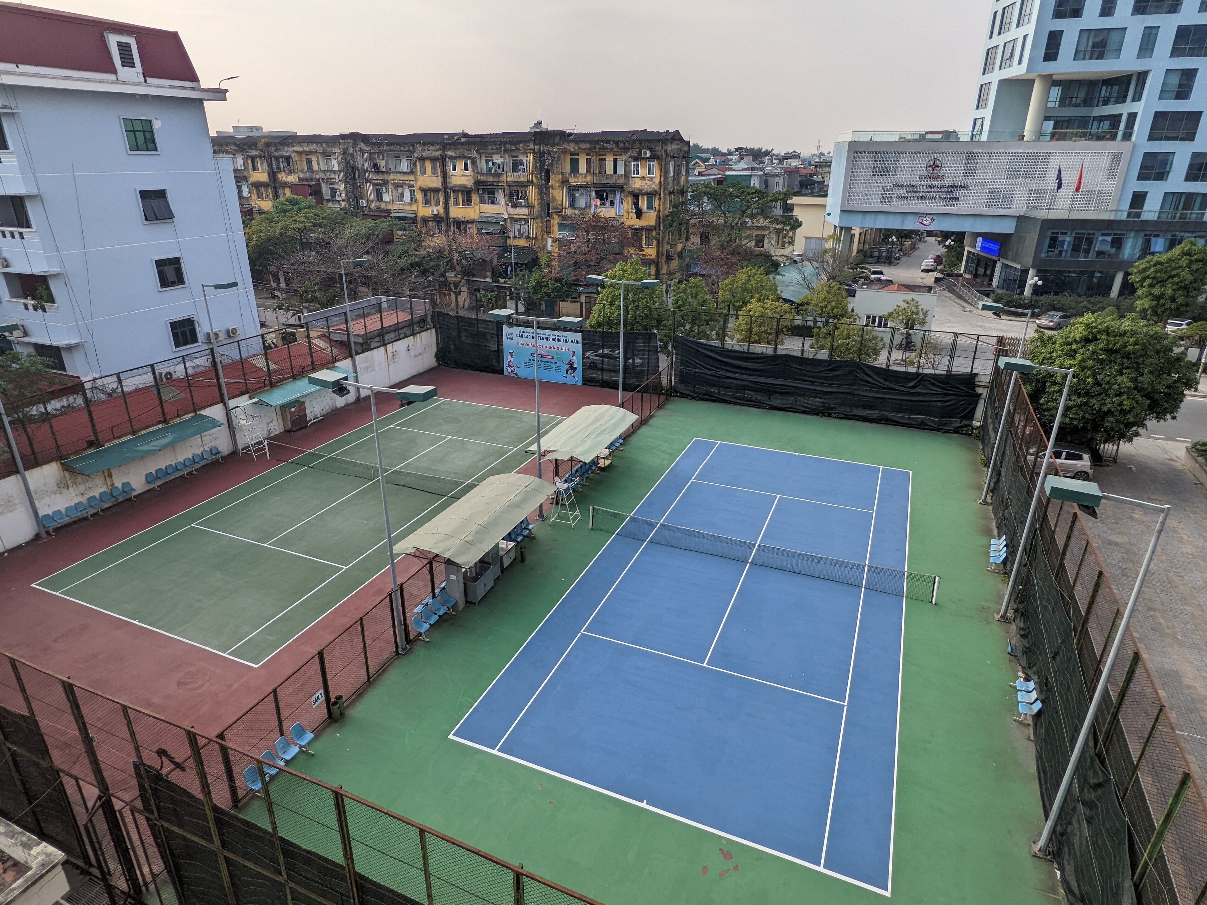 Una cancha de tenis al aire libre con dos pistas, rodeada por una cerca y adyacente a edificios residenciales.