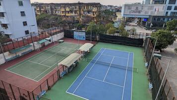 Una cancha de tenis al aire libre con dos pistas, rodeada por una cerca y adyacente a edificios residenciales.