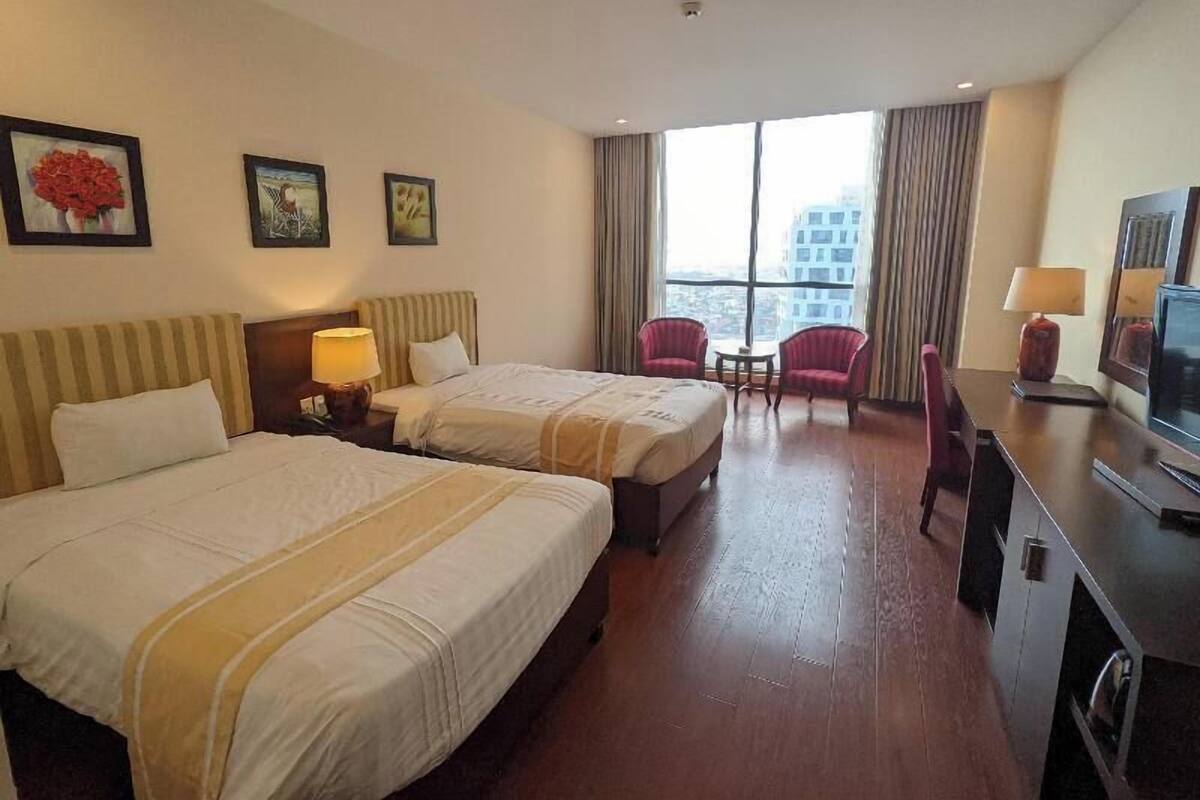 A hotel room with two beds, a TV, a desk, and a view of buildings through large windows.