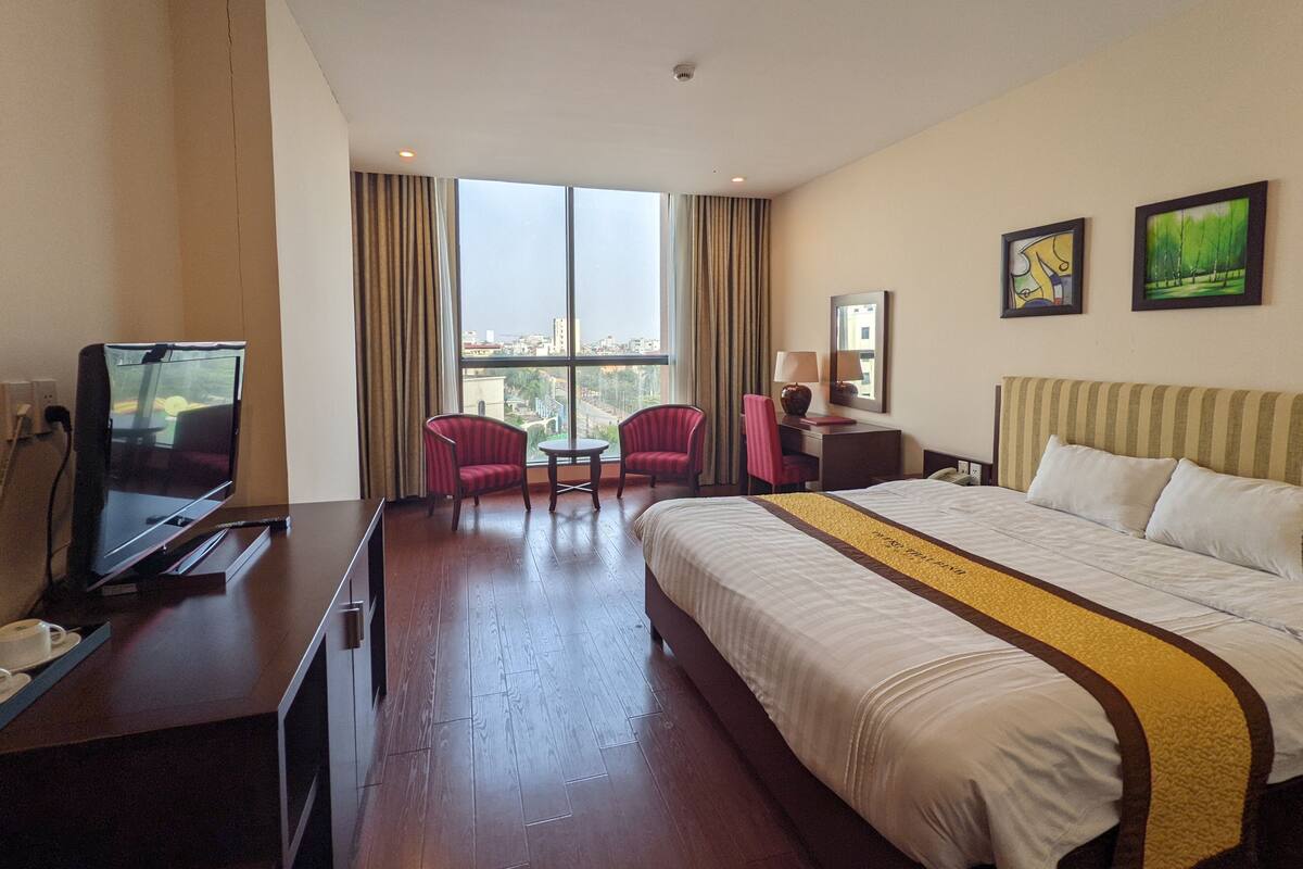 A hotel room with a large bed, a desk with a television, two armchairs, a small table, and a view of the city through large windows.