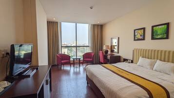 Una habitación de hotel con una cama grande, un escritorio con televisor, dos sillones, una mesita y vistas a la ciudad a través de amplias ventanas.