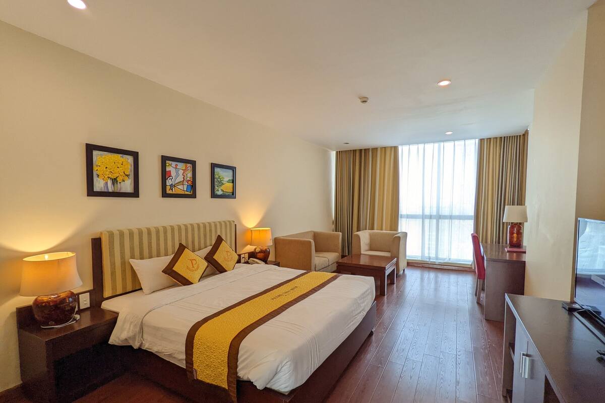 A hotel room with a large bed, two armchairs, a desk, and a TV. There are three framed pictures on the wall, a window with curtains, and a lamp on the desk.