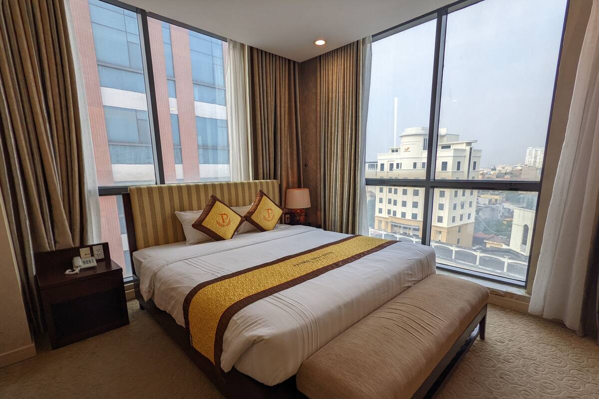 A hotel room with a large bed, a bedside table with a phone, and a view of the city through large windows.