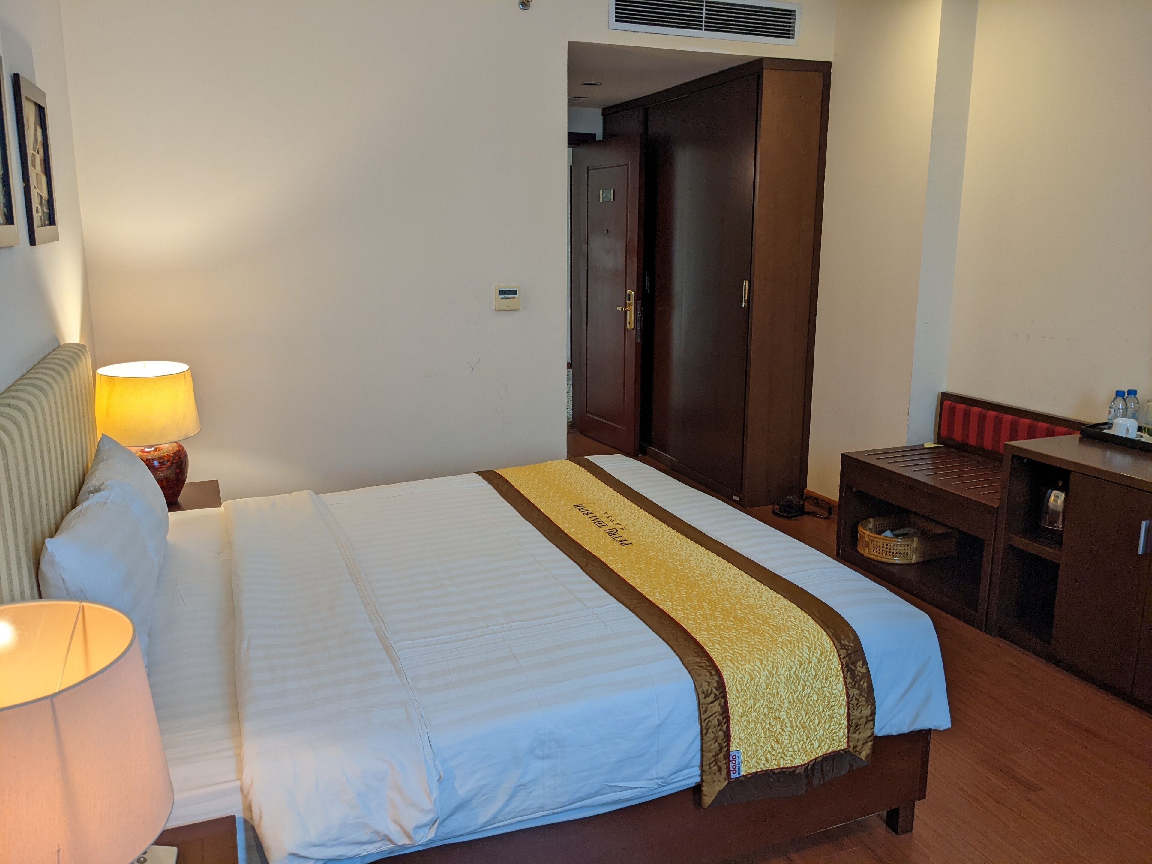 A hotel room with a bed, a nightstand with a lamp, a wooden wardrobe, and a small table with a tray.