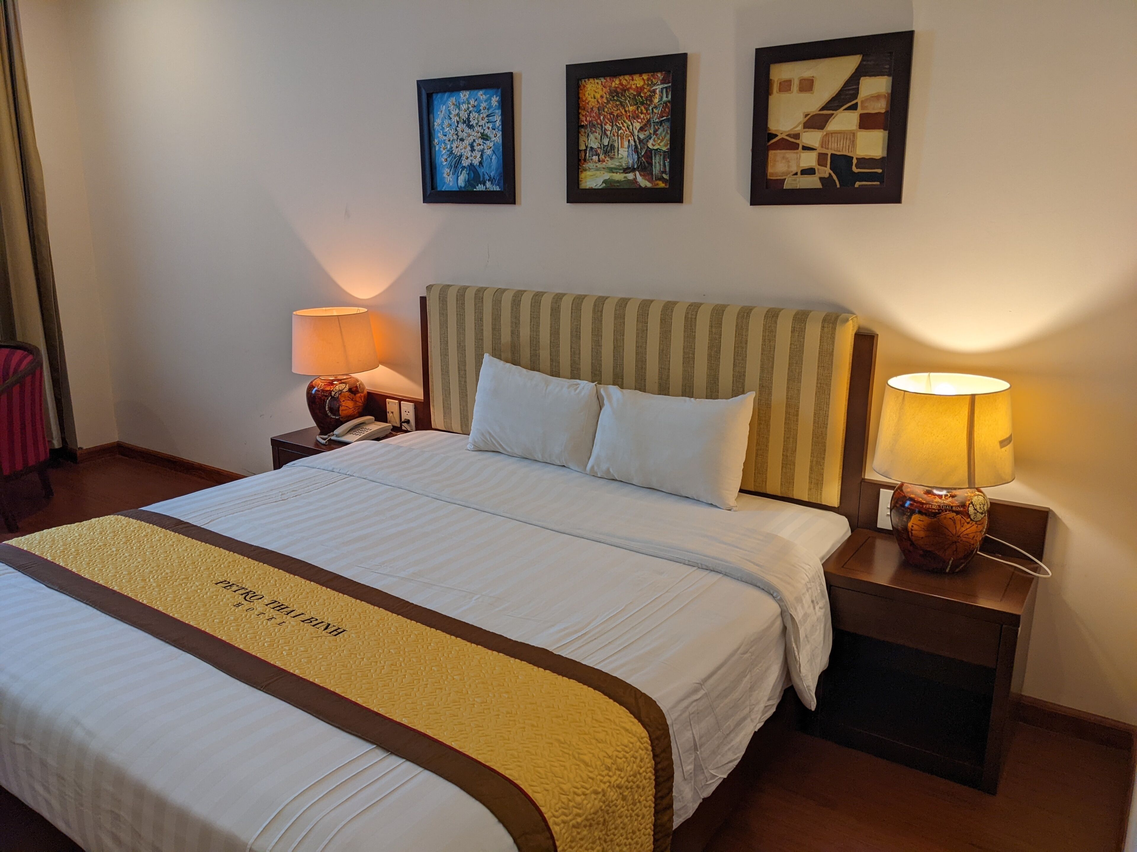 A hotel room with a large bed, two bedside lamps, a telephone, and three framed artworks on the wall.