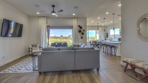 Un'ampia zona giorno con un divano grigio, una TV a schermo piatto montata a parete e un'area pranzo con una cucina sullo sfondo.