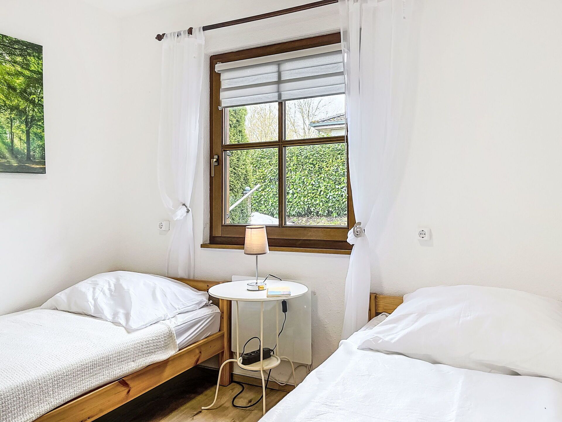 A small room with two single beds, a window with a view of greenery, a small table with a lamp, and a power outlet.