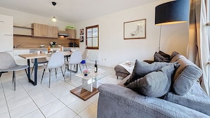 A modern living room with a grey sofa, a dining table with chairs, a kitchen area, and a large window with curtains. - Cosy apartment in villa for 6 guests with WIFI, TV, terrace and pets allowed (Bad Wildungen)