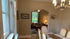 A dining room with a wooden table, chairs, and a chandelier. There is a window on the left and a door leading to another room.