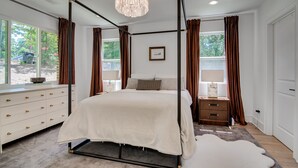 Une chambre à coucher avec un lit à baldaquin, une commode blanche, une table de chevet, un lustre et une vue sur l’extérieur.