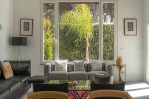 A living room with a sofa, a coffee table, and a TV. There are large windows with a view of greenery outside. - LV Premier LA2 heated pool, AC, garden, sea view (Sesimbra)