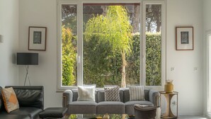 Una sala de estar con un sofá, una mesa de centro y un televisor. Amplias ventanas ofrecen vista a zonas verdes.