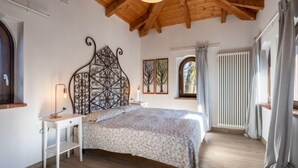 A bedroom with a wooden ceiling, a bed with a floral bedspread, a nightstand with a lamp, a window with curtains, and a radiator. - La Quercia by Interhome (Mondovì)