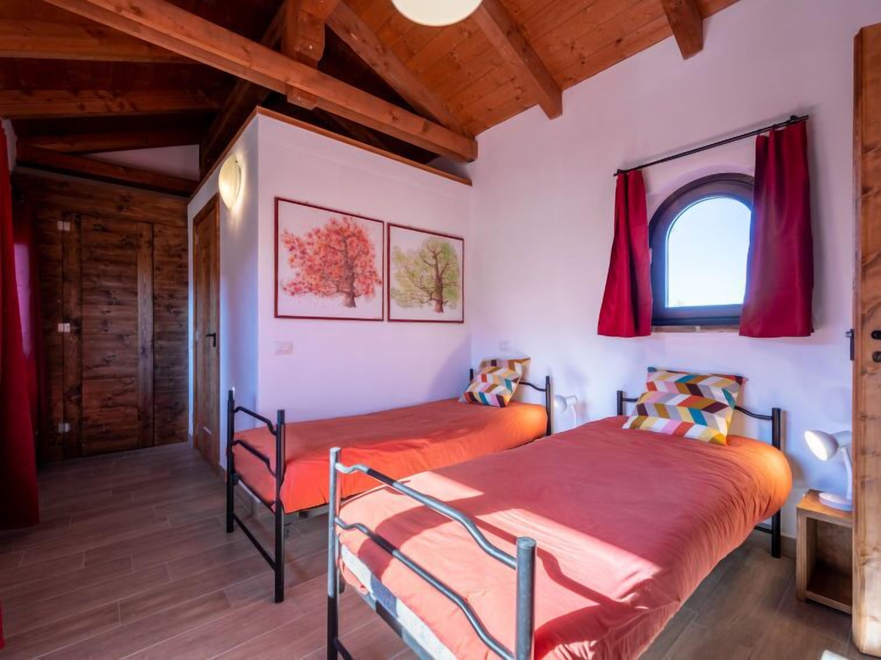 Una stanza con due letti a castello, travi in legno al soffitto, una finestra con tende rosse e due quadri appesi al muro.