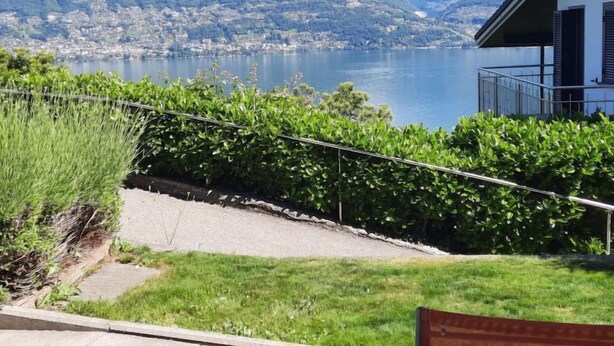 Une vue sur un lac, avec des montagnes en arrière-plan, un bâtiment doté d’un balcon et une zone herbeuse délimitée par une clôture.