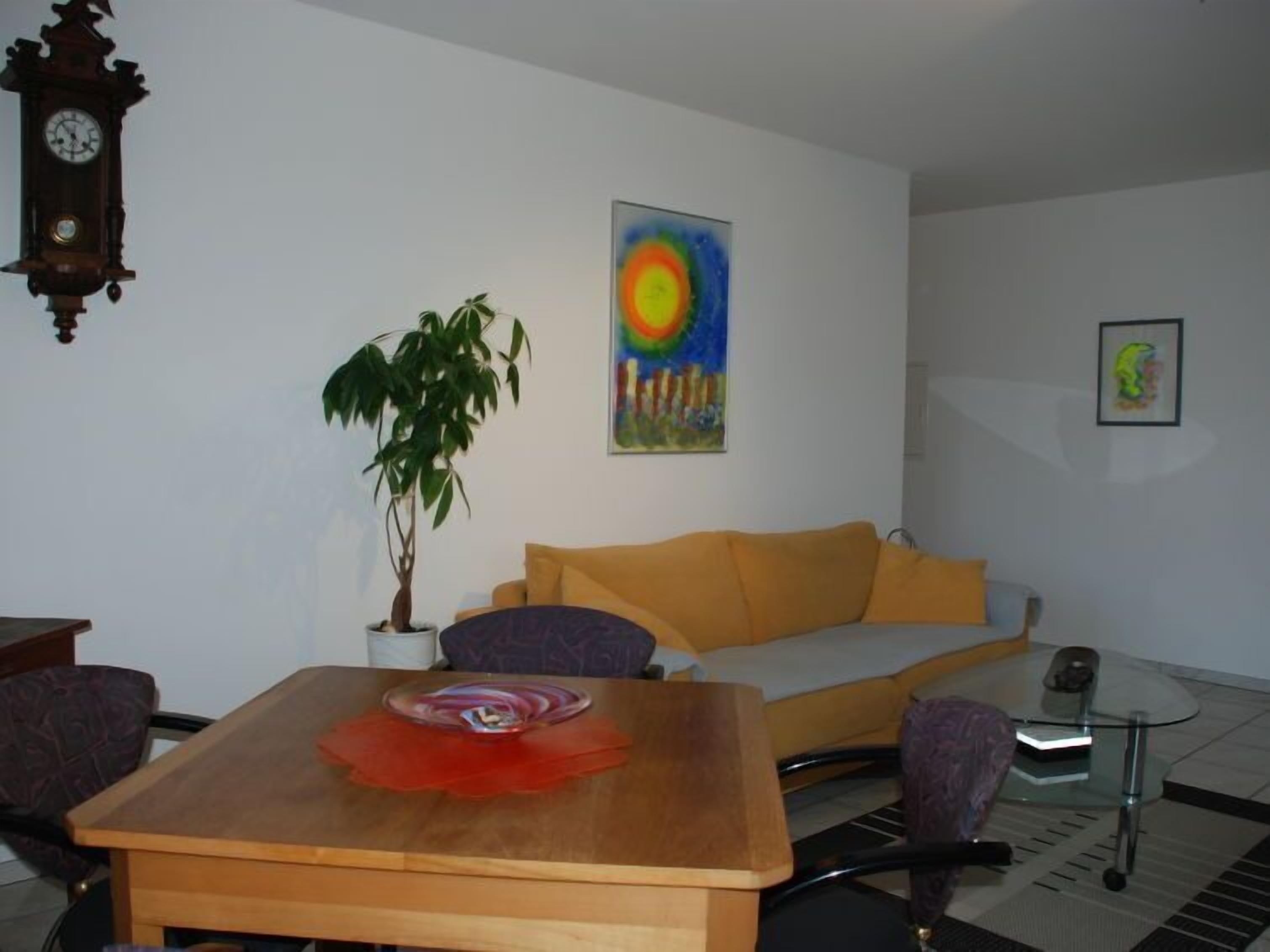 A living room with a wooden table, a sofa, a potted plant, a painting, and a clock on the wall.
