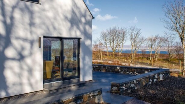 Et moderne hus med en stor glasdør, sten trapper og udsigt over et landligt landskab med træer og et hegn.