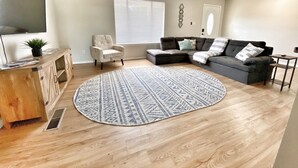 A living room with a wooden floor, a grey sectional sofa, a white armchair, a round patterned rug, a ceiling fan, and a window with blinds. - Game Room - Mountain Views - King Bed - near Golf Course - Intel - Rust Medical  (Albuquerque)