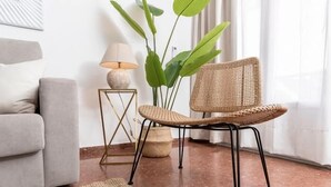 Un salon avec un canapé gris, un fauteuil en osier, une petite table d’appoint avec une lampe et une grande plante verte.