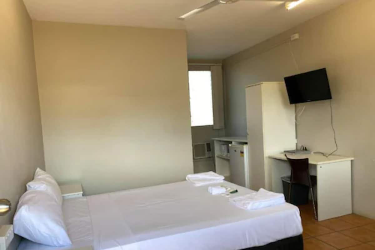 A bedroom with a bed, a desk, a chair, a TV, and a window.